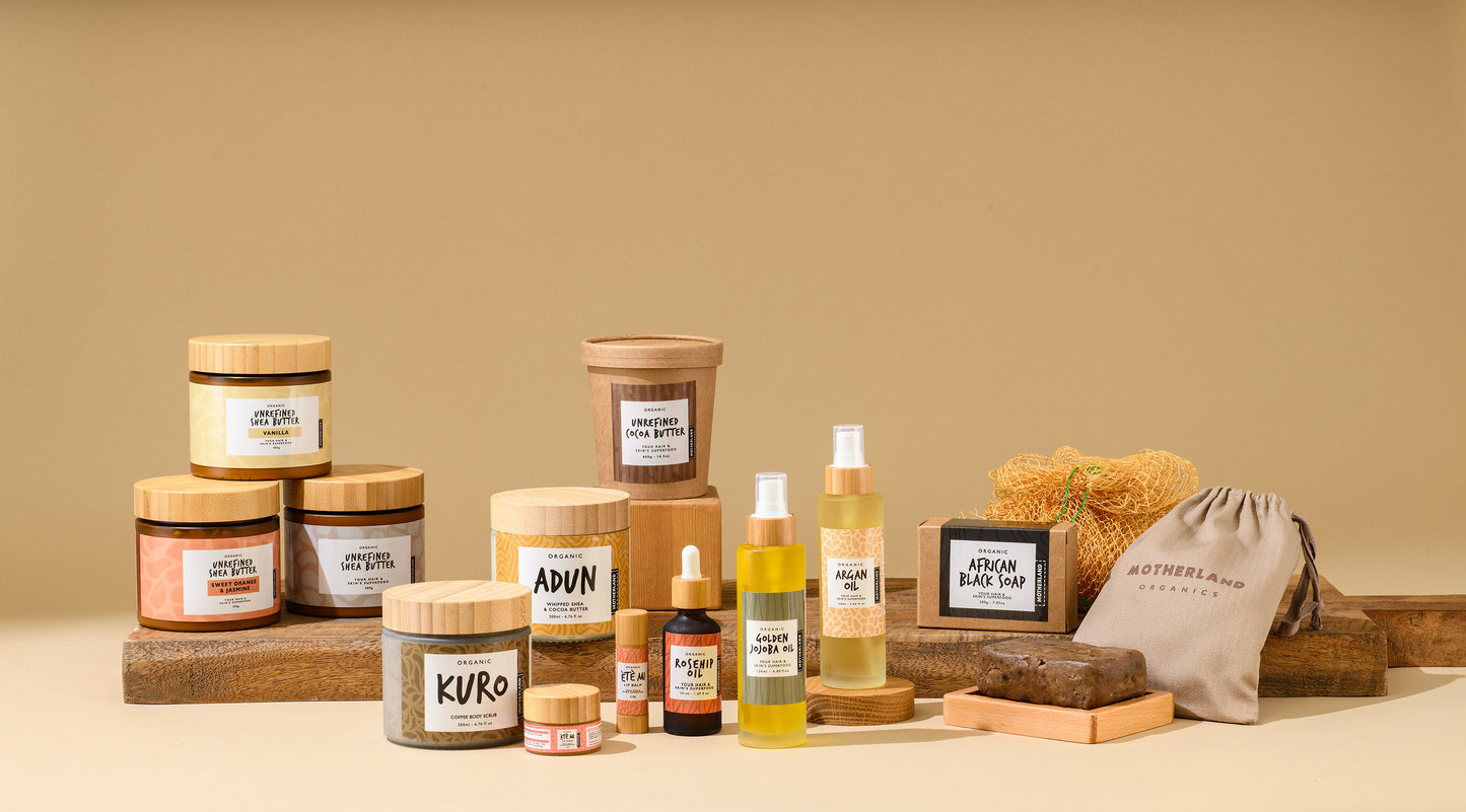 A wider shot of a curated collection of  Motherland Organics products featuring African black bar soap, unrefined shea butter, whipped shea body butter, raw cocoa butter, organic lip balm and lip scrub, cold-pressed rosehip oil, argan oil, golden jojoba oil, and a coffee body scrub — natural, vegan skincare for healthy, nourished skin, lips, and hair.