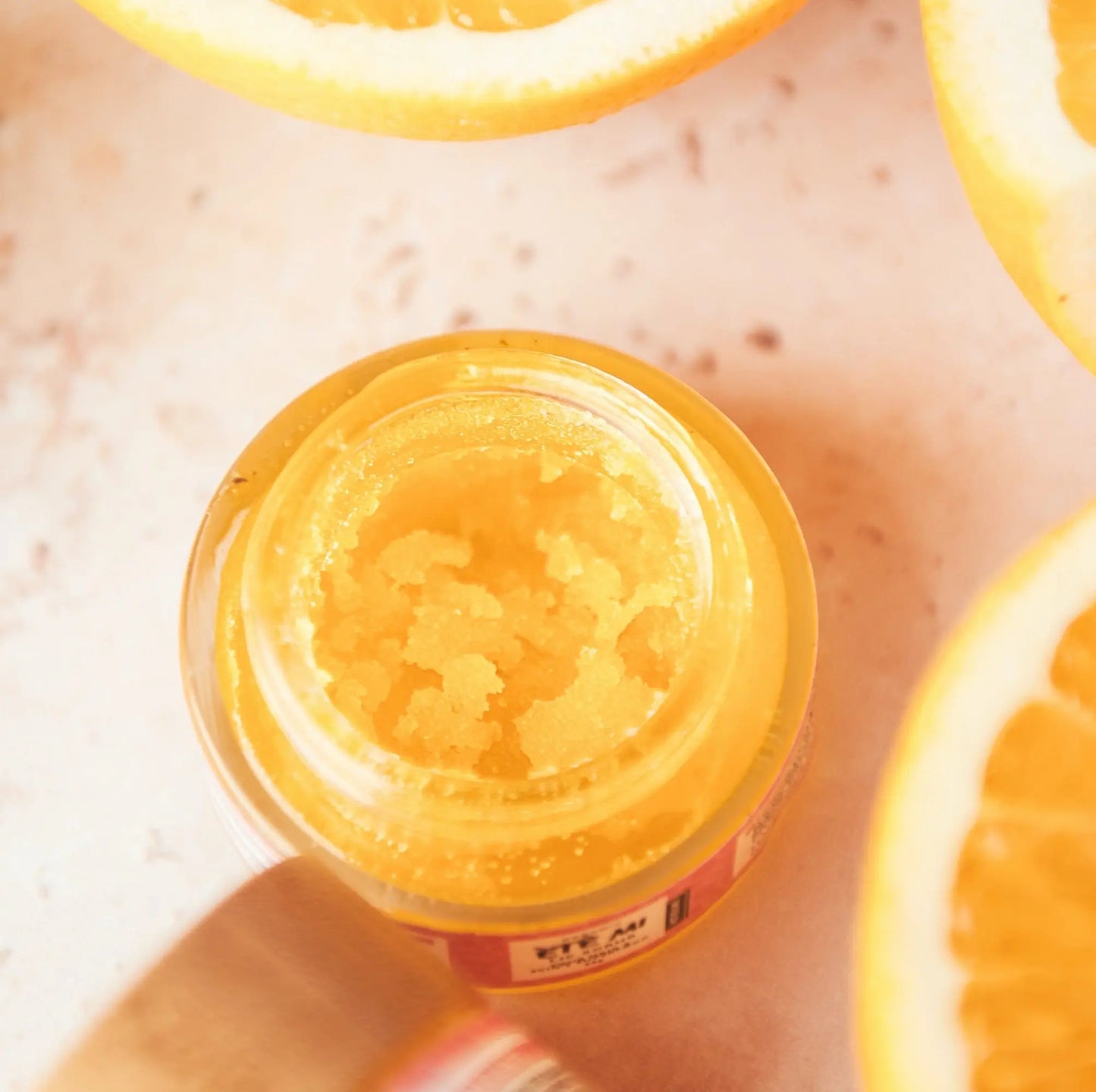 Bright citrus lip scrub in a glass jar, shown with fresh orange slices. Exfoliating lip scrub made with natural ingredients to smooth and soften dry, chapped lips.