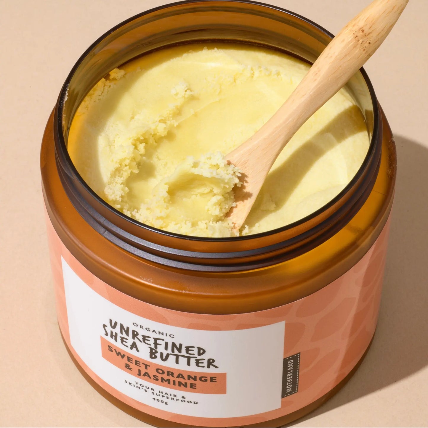 Open jar of Motherland Organics Unrefined Shea Butter – Sweet Orange & Jasmine, with a wooden spoon scooping the firm, natural texture of raw shea butter. This organic body butter works as a shea butter hair butter, moisturiser for dry skin, and shea butter for eczema. Made with pure, unrefined African shea butter, it's ideal as a shea butter face cream, hand cream, or lip balm. Rich in nutrients, this shea butter cream is vegan, natural, and perfect for full-body use — from skin to scalp.