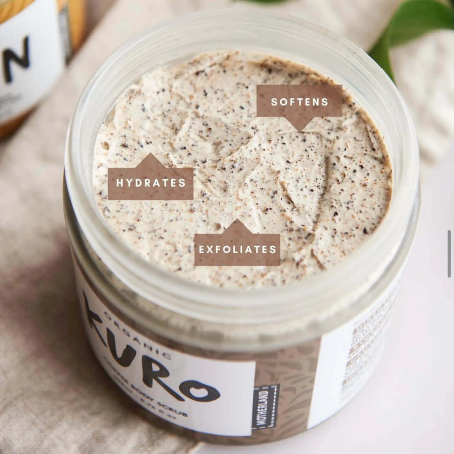 Open jar of KURO coffee scrub showing whipped texture with labels highlighting exfoliates, hydrates, and softens — organic body scrub made with coffee grounds for smooth, radiant skin.
